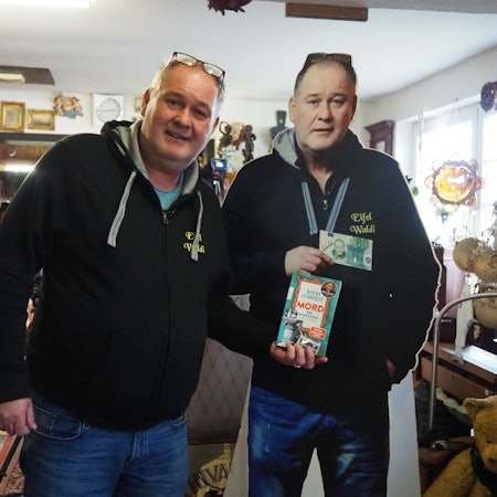 Ein Mann im Pullover und mit einer Brille, die auf seinem Kopf aufliegt, steht neben einem Pappaufsteller von sich und hält ein Buch in die Kamera.