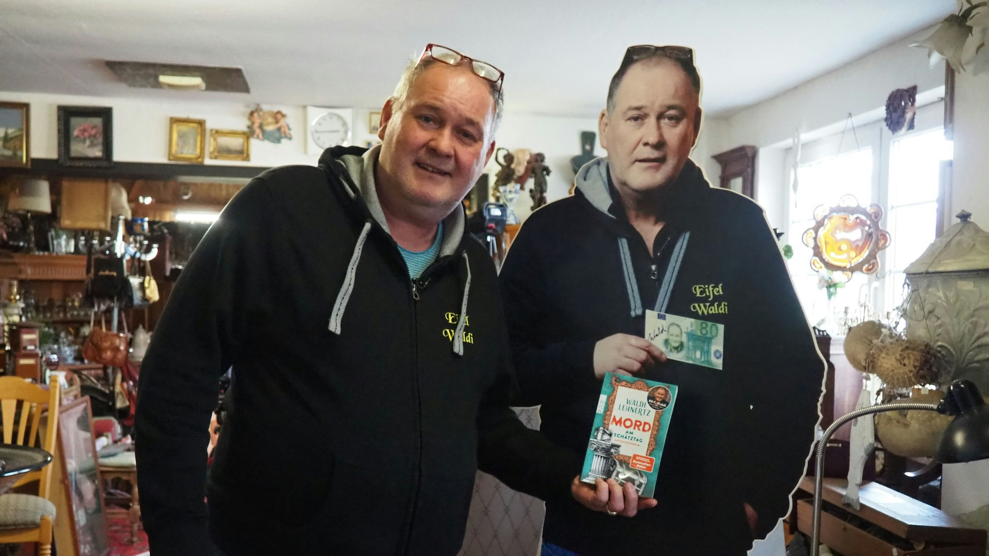 Ein Mann im Pullover und mit einer Brille, die auf seinem Kopf aufliegt, steht neben einem Pappaufsteller von sich und hält ein Buch in die Kamera.