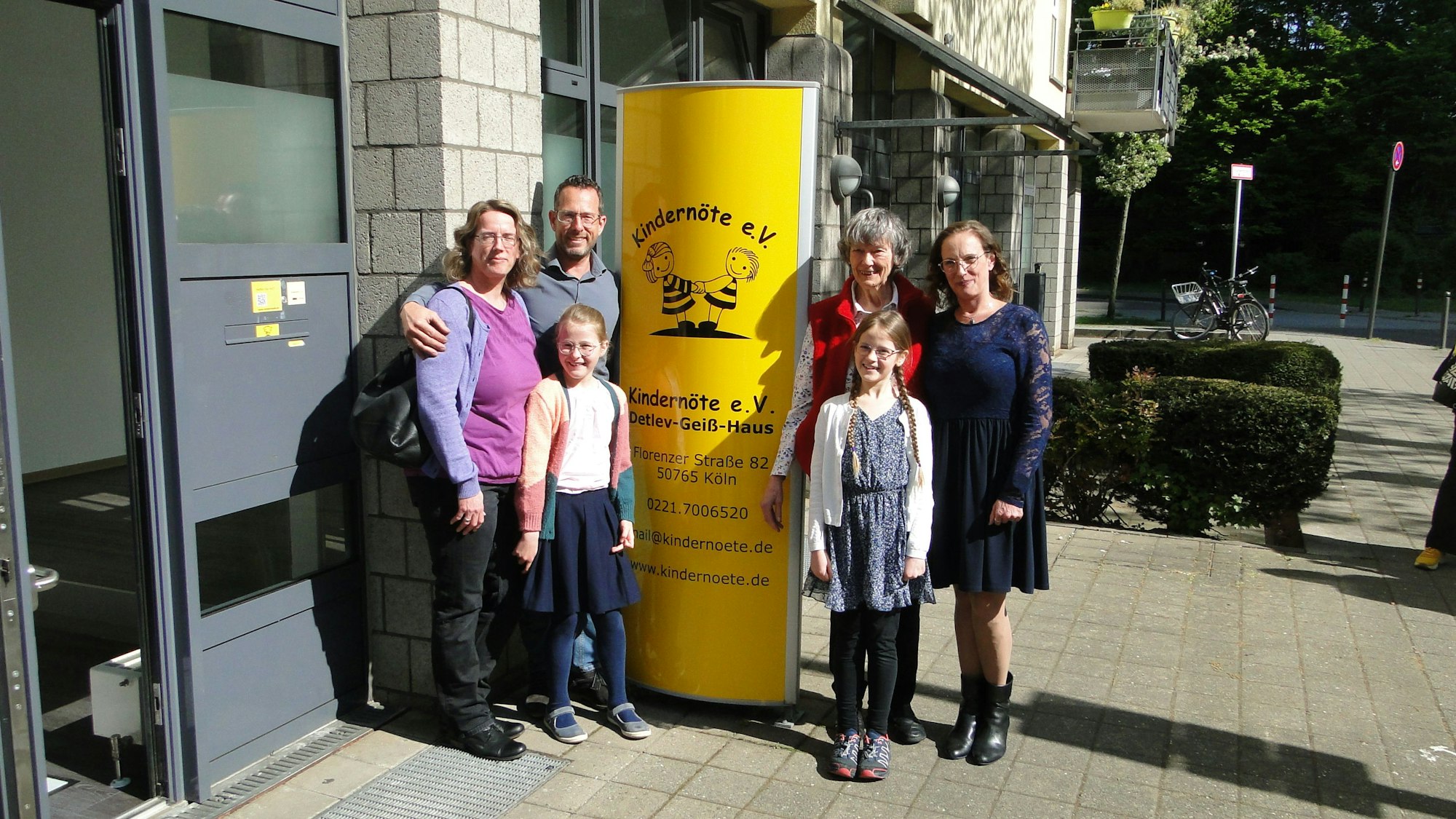 Vier Erwachsene und zwei Kinder stehen vor einem Hauseingang links und rechtrs von einer gelben Säule mit den Informationen zum neuen Detlev-Geiß-Haus.
