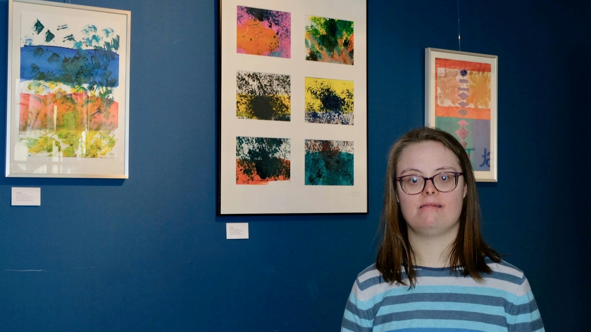 Eine junge Frau mit schulterlangem Haar und Brille steht vor einer blauen Wand, an der drei abstrakte Bilder in Pastellfarben hängen.
