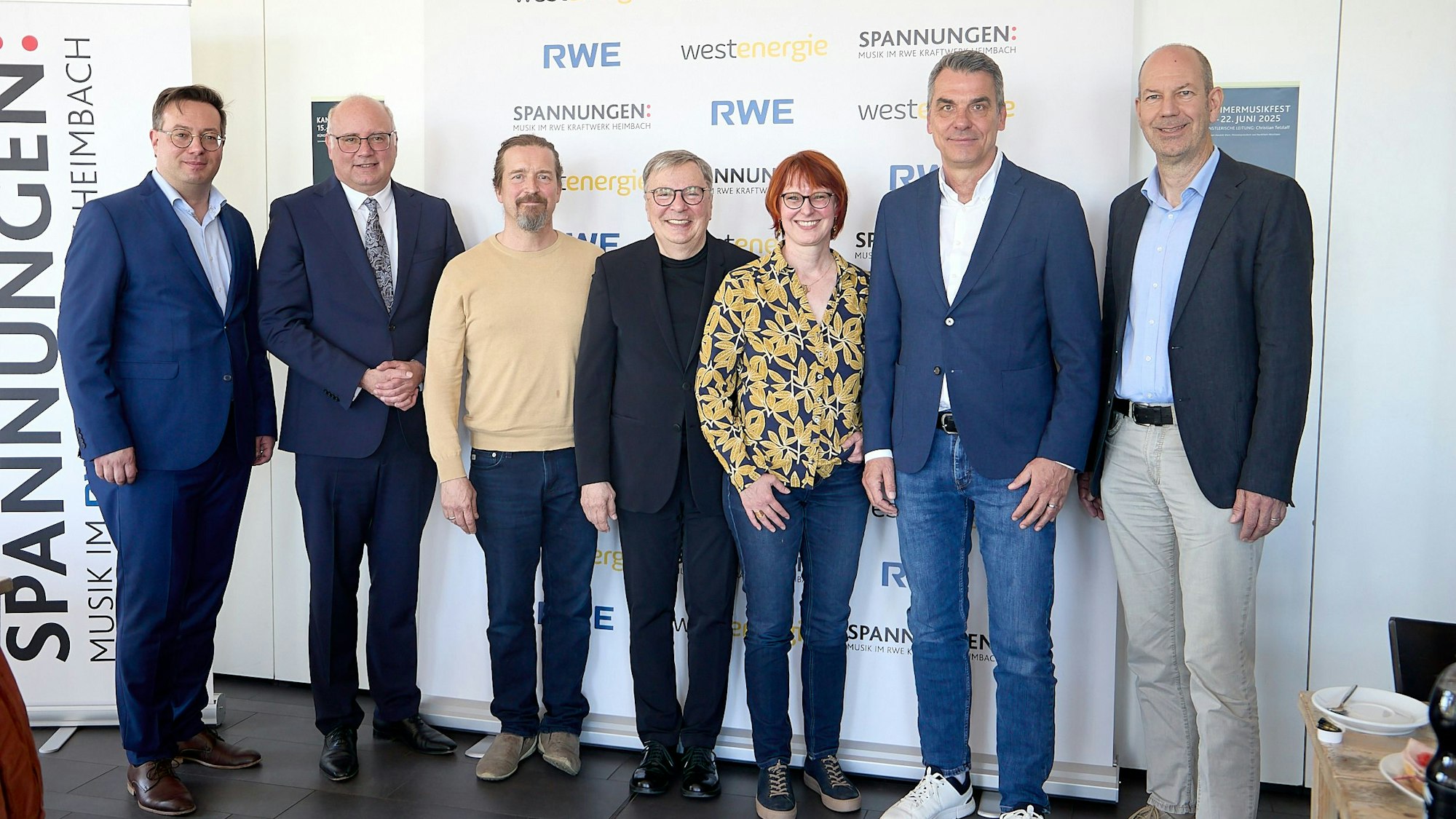 Sieben Menschen stehen in einer Reihe. Links ist ein Plakat des Festivals Spannungen zu sehen.