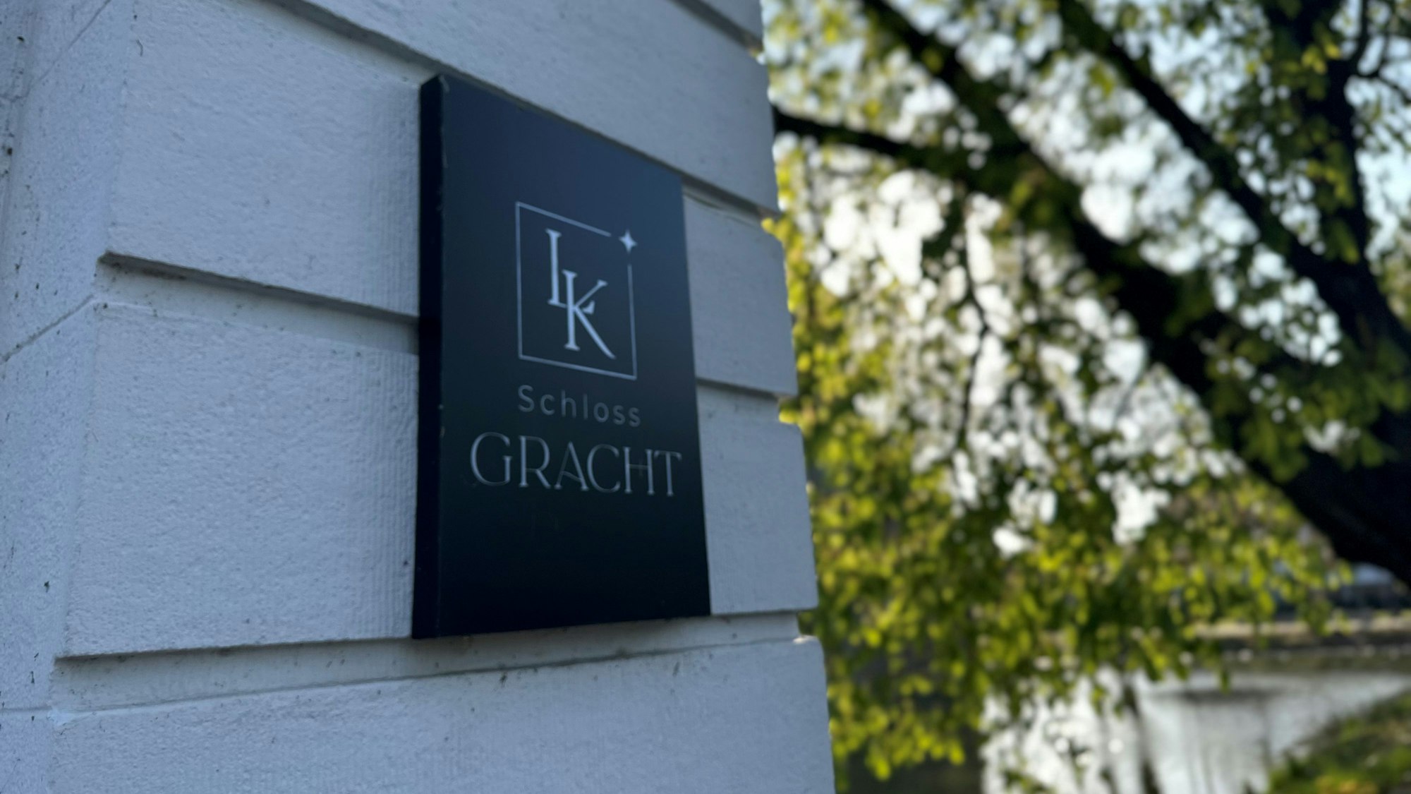 Zu sehen ist ein schwarzes Schild auf einem weißen Pfeiler mit der Aufschrift Schloss Gracht.