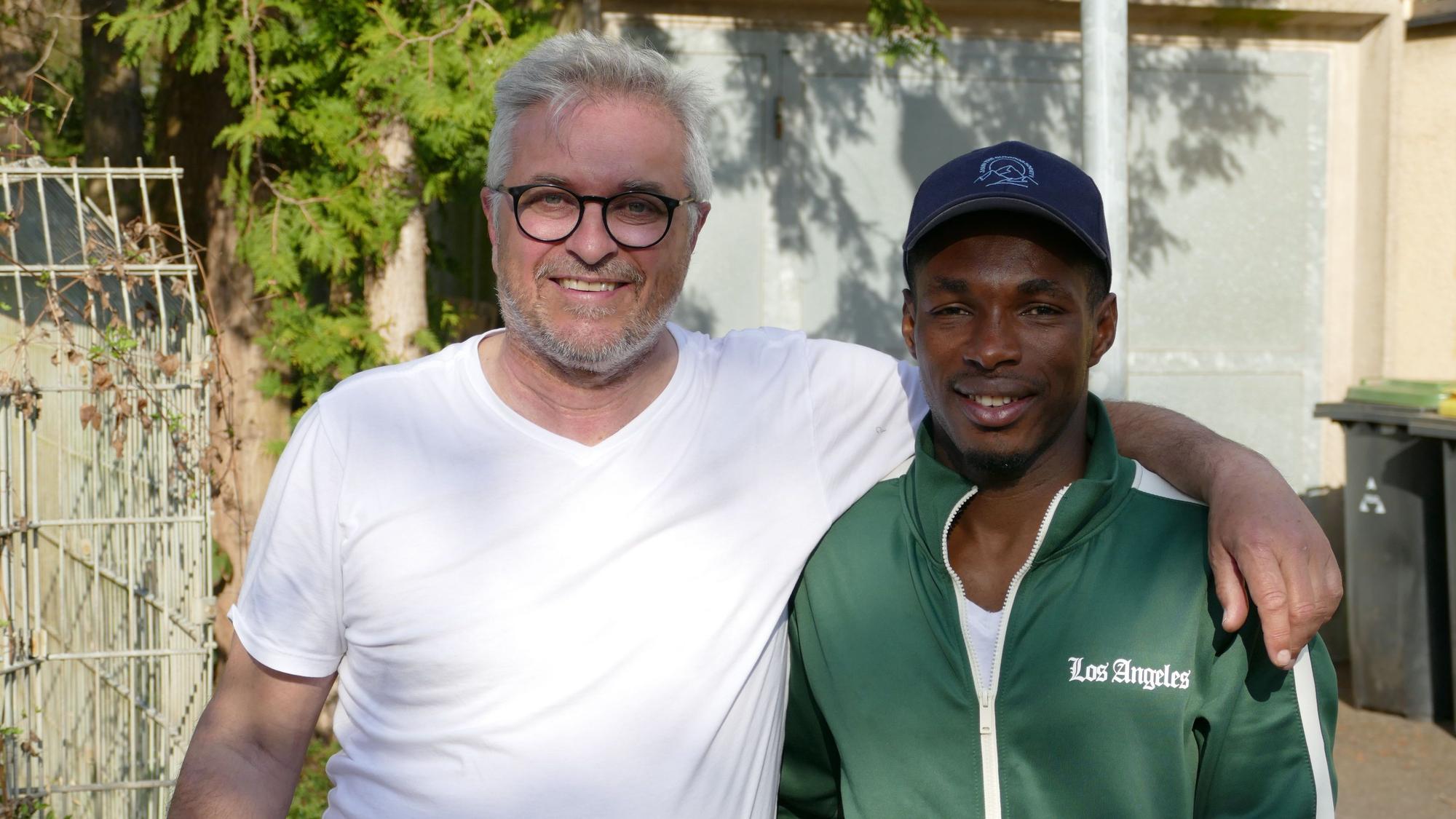 Ein Mann im weißen T-Shirt, mit Brille und grauen Haaren, steht neben einem dunkelhäutigen Mann mit Baseballkappe und grüner Trainingsjacke.