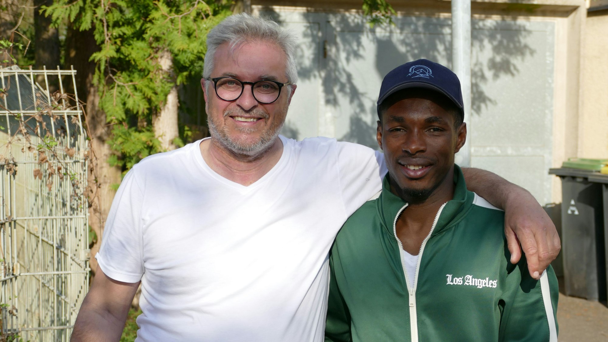 Ein Mann im weißen T-Shirt, mit Brille und grauen Haaren, steht neben einem dunkelhäutigen Mann mit Baseballkappe und grüner Trainingsjacke.