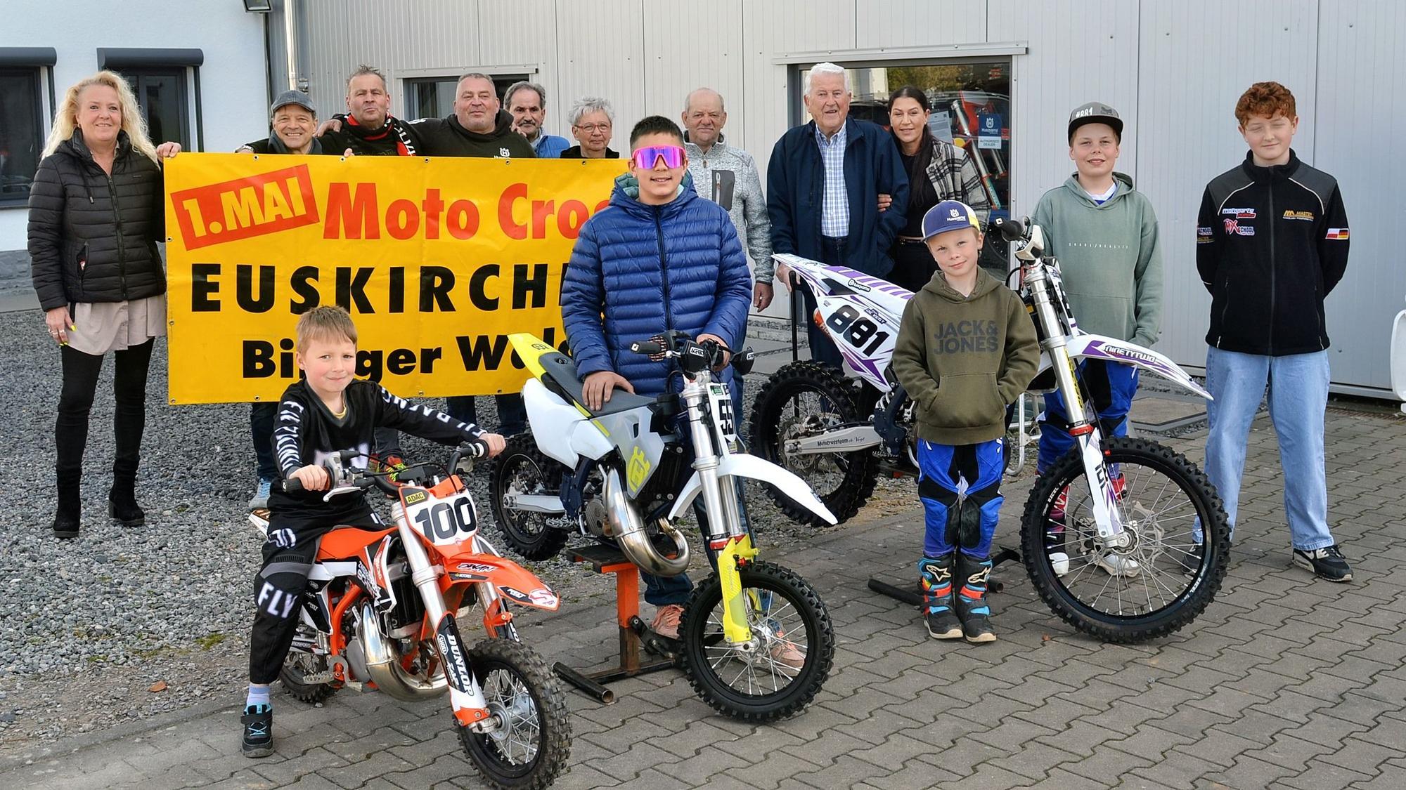 Fünf Kinder und Jugendliche stehen an Motocross-Maschinen, dahinter einige Erwachsene, die mit einem Plakat auf den Wettbewerb am 1. Mai im Billiger Wald hinweisen.