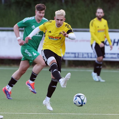 Das Foto zeigt einen Spieler des VfR Bachem im gelben Trikot, der von zwei Horremern in grünen Trikots am Abspiel nicht gehindert werden kann.