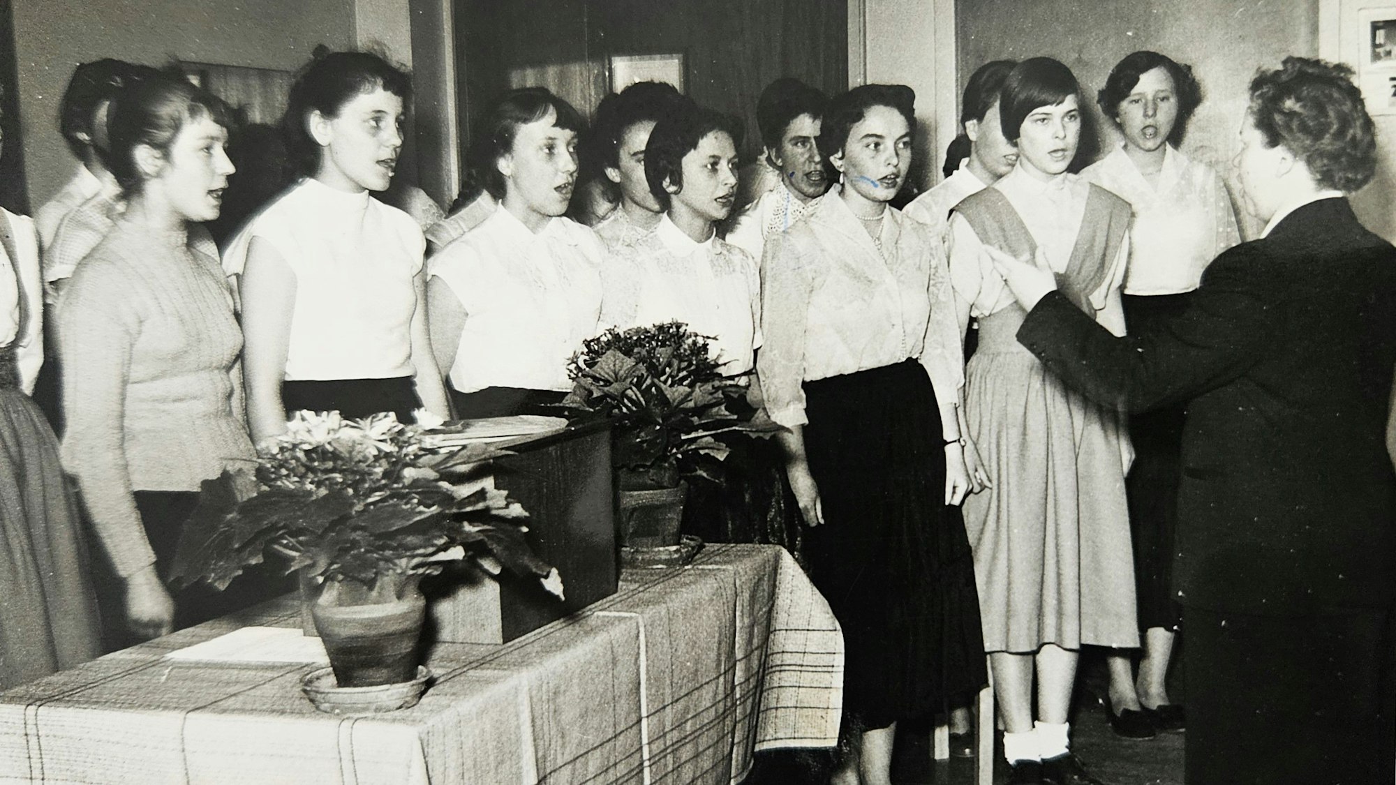 Mädchen stehen nebeneinander und singen. Vor ihnen steht eine Lehrerin und dirigiert.
