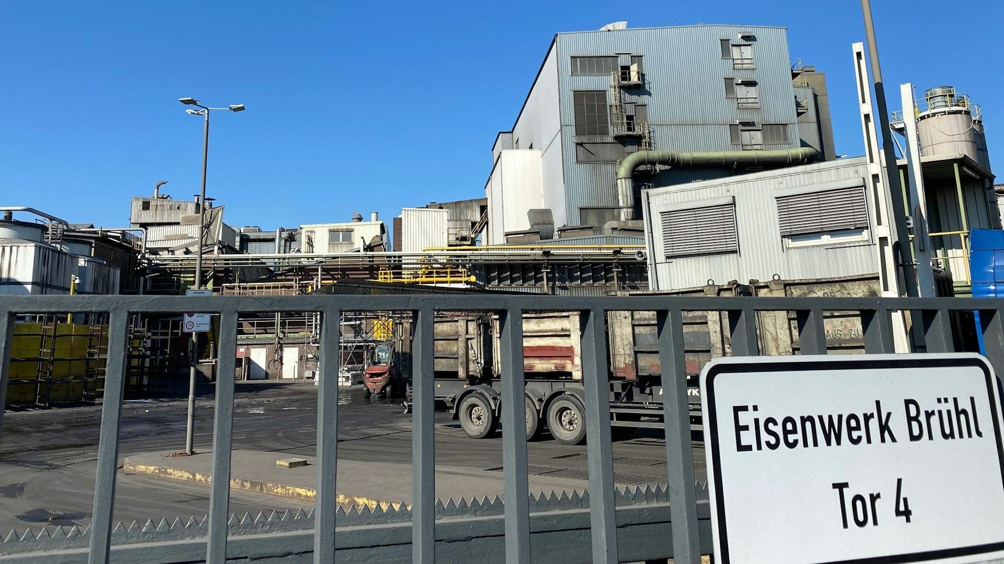 Zu sehen ist ein Zaun am Werksgelände des Eisenwerks und dahinter die Werkshallen.