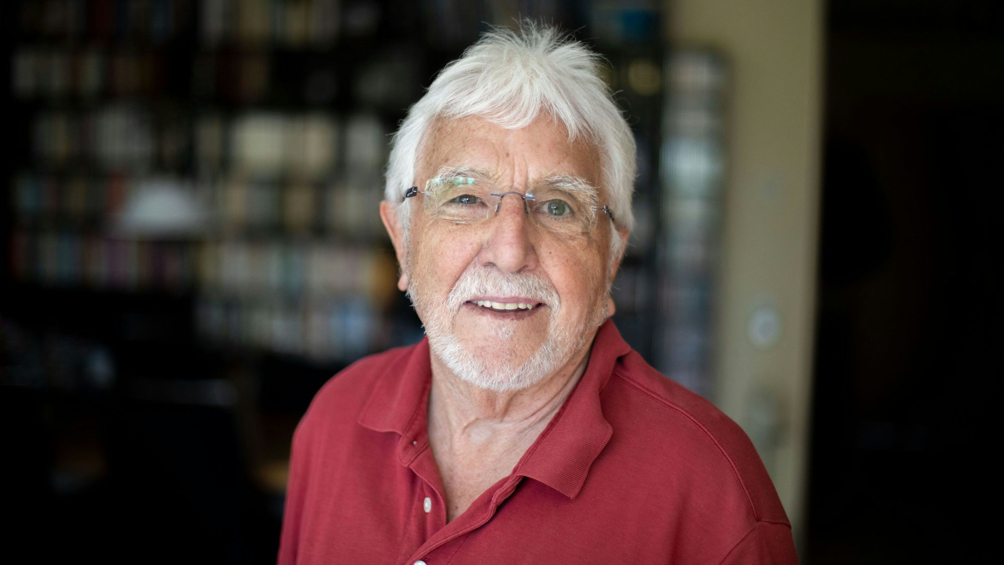 Ein Mann mit weißem Haar und weißem Bart steht vor einer Schrankwand.