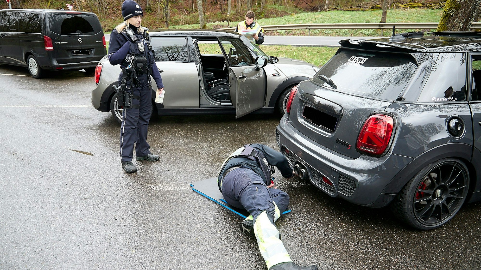 Mehrere Fahrzeuge werden auf einem Parkplatz von Polizisten inspiziert. Einer der Beamten liegt am Boden und schaut unter das Auto. Eine Polizistin schaut dabei zu.
