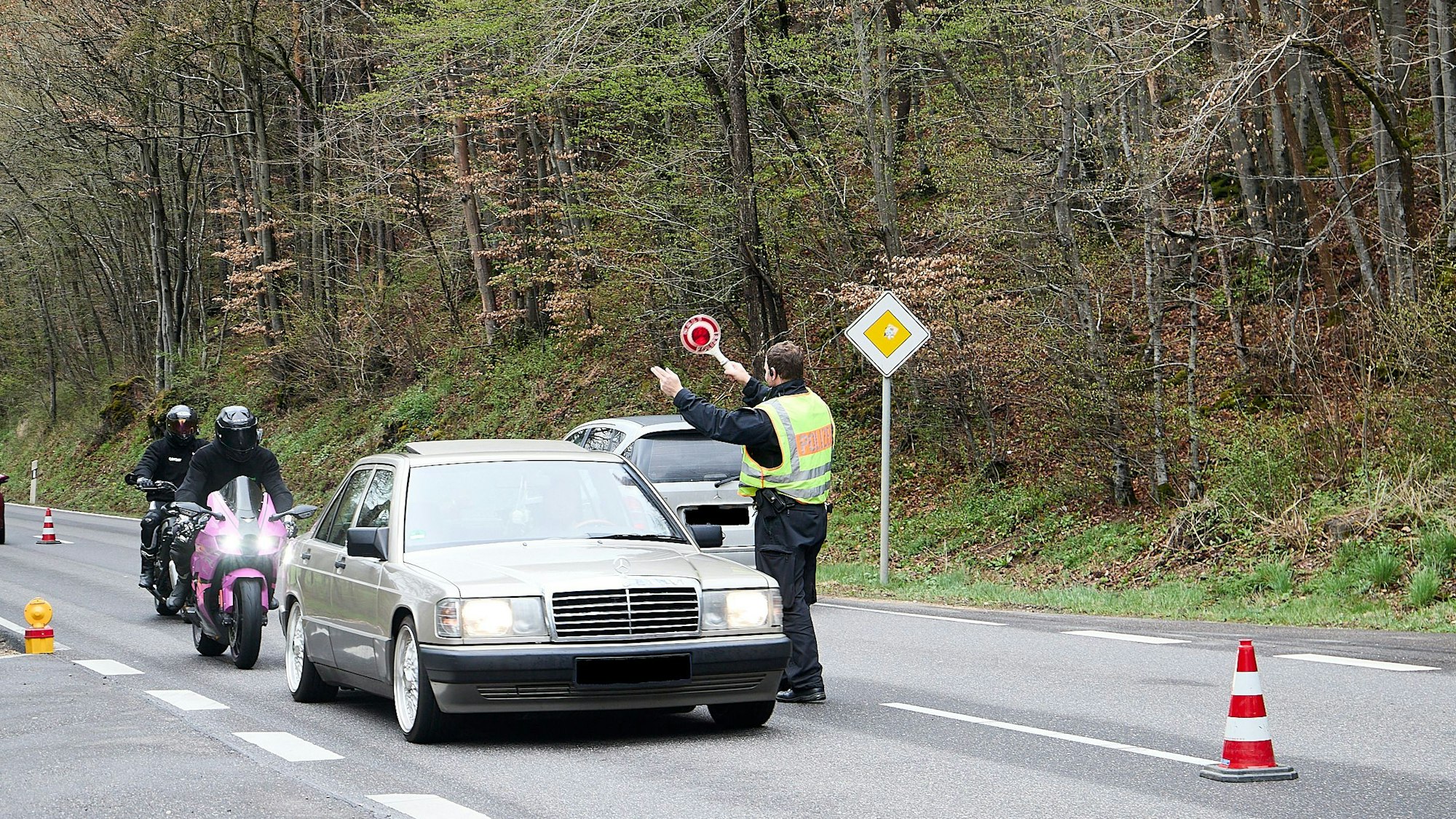 Ein Polizist steht auf der Straße, lässt einen Mercedes passieren und winkt mit der Kelle zwei nachfolgende Motorradfahrer heraus.