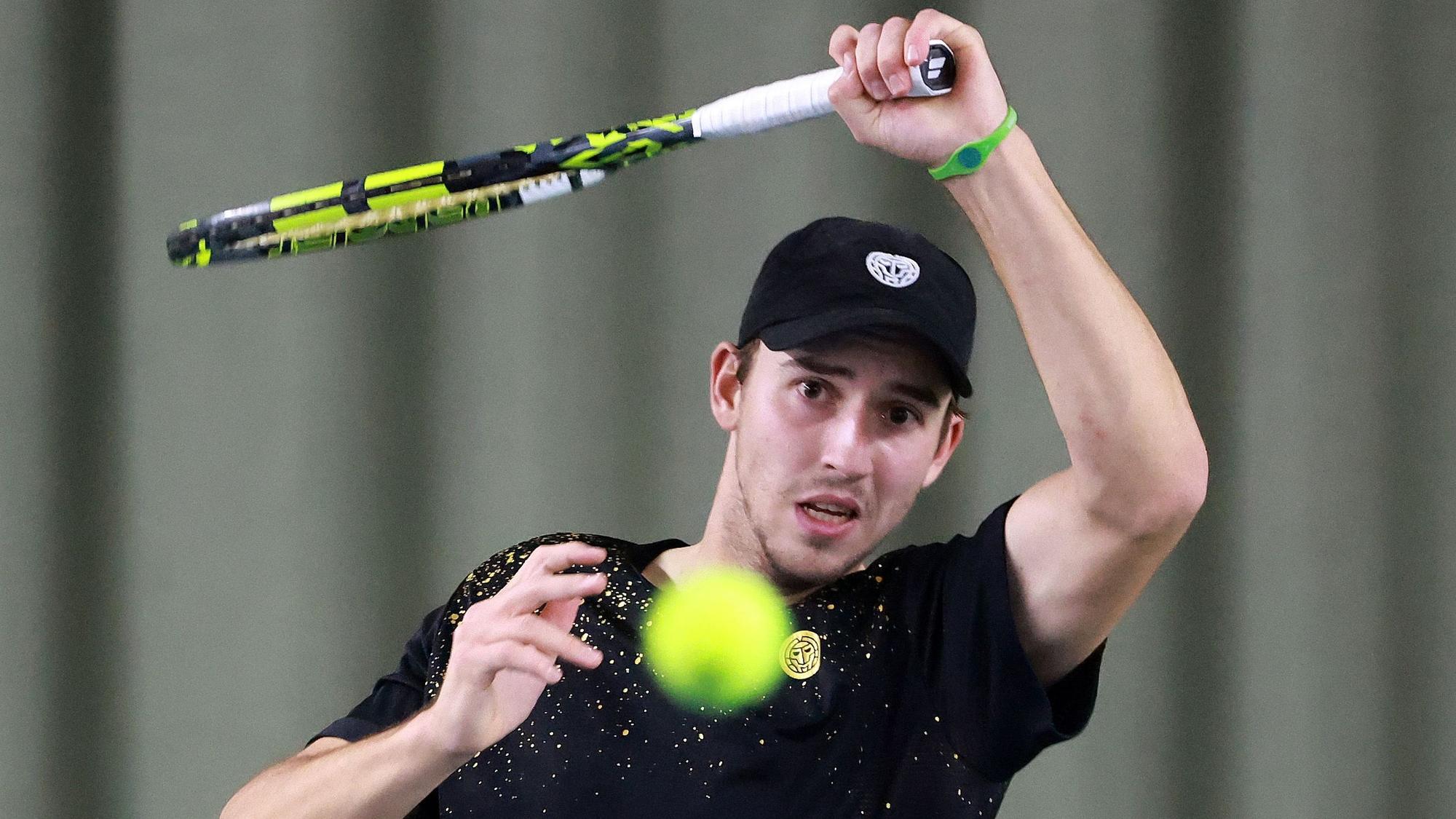 Das Foto zeigt einen Tennisspieler mit schwarzem T-Shirt und schwarzer Kappe beim Return.