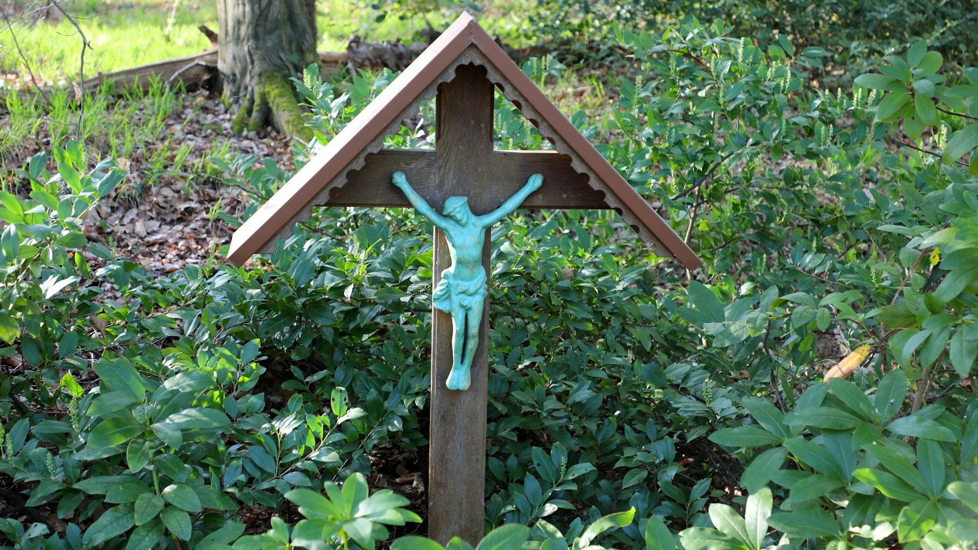 Ein Wegekreuz steht zwischen Grünpflanzen am Waldrand.