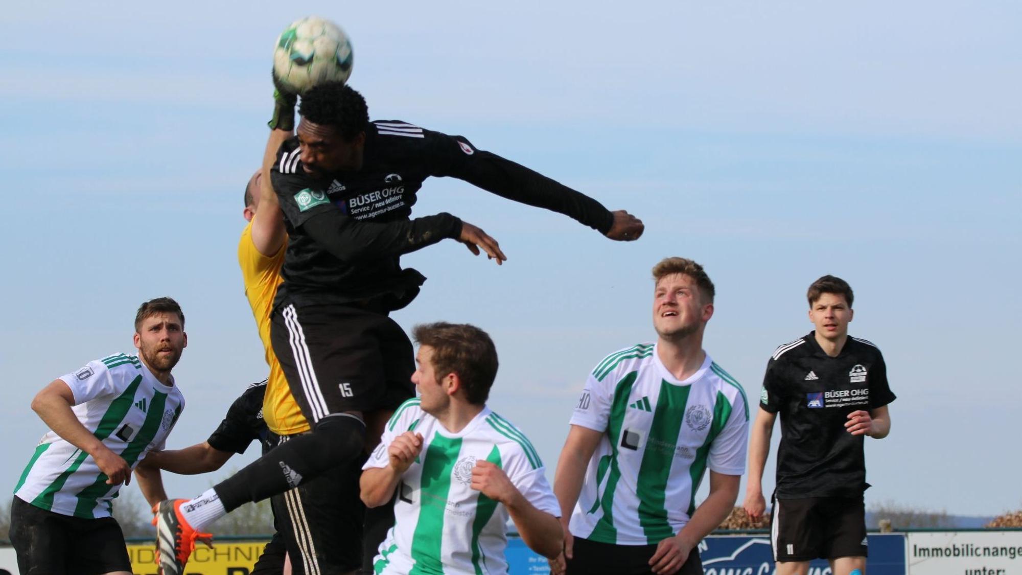 Luftkampf zwischen Schöneseiffens Torwart und Nierfeld Verteidiger Smart, der hochspringt, aber den Ball, den der Torwart zu fassen bekommt, nicht richtig trifft. Vier weitere Spieler schauen zu.