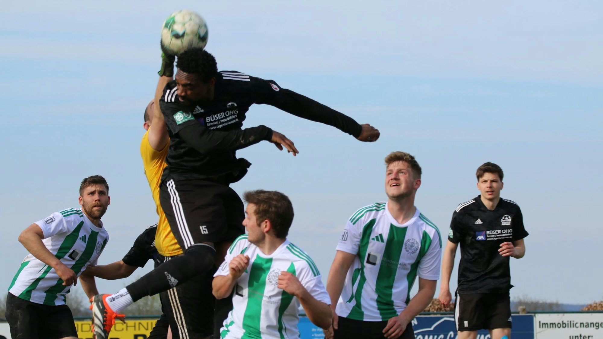 Luftkampf zwischen Schöneseiffens Torwart und Nierfeld Verteidiger Smart, der hochspringt, aber den Ball, den der Torwart zu fassen bekommt, nicht richtig trifft. Vier weitere Spieler schauen zu.