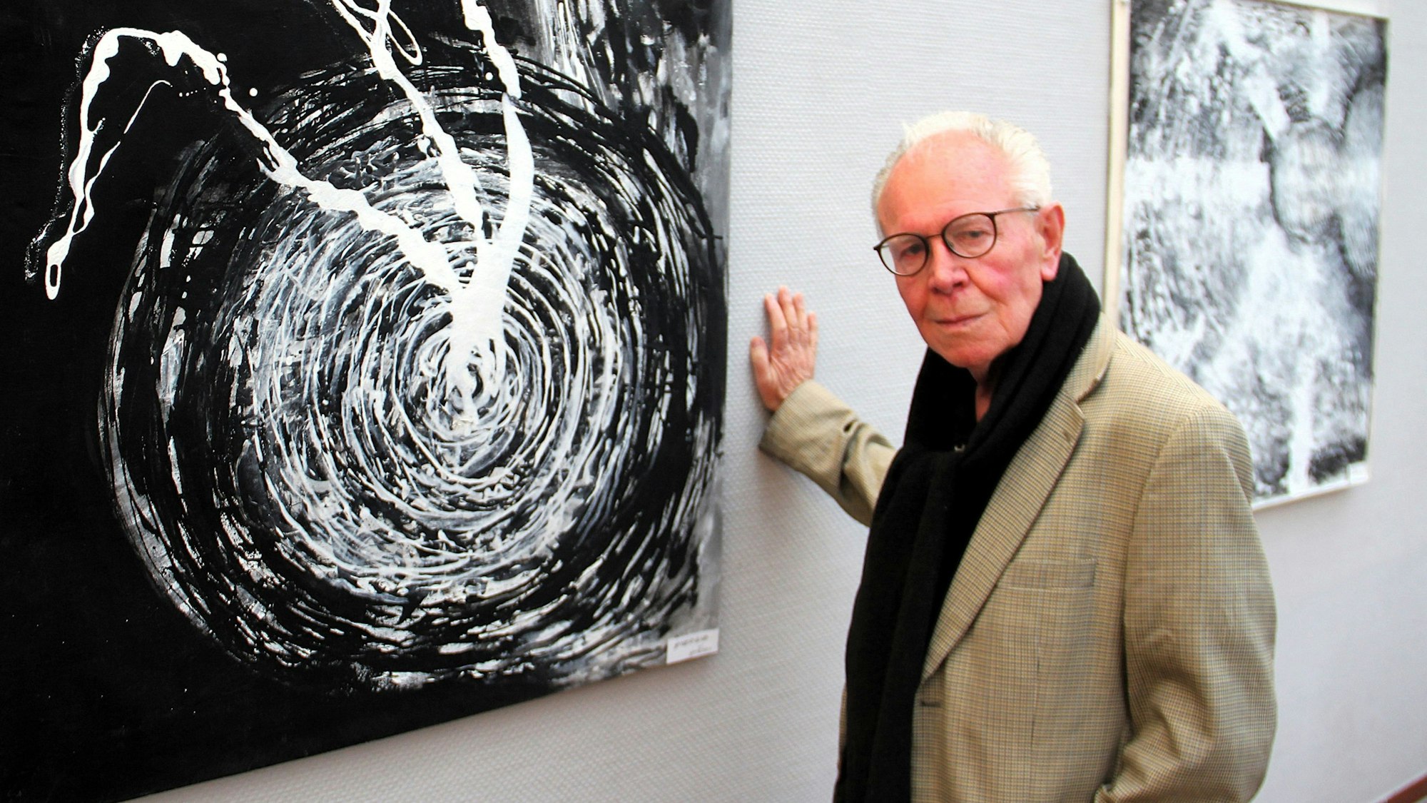 Ein älterer Herr steht in einer Galerie neben seinem abstrakten Schwarz-Weiß-Werk.