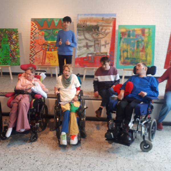 Menschen, teils im Rollstuhl sitzen vor einer Bühne, auf der fünf Bilder auf Staffleien stehen. Der Künstler, Mikail Akar, steht vor seinen Bildern.