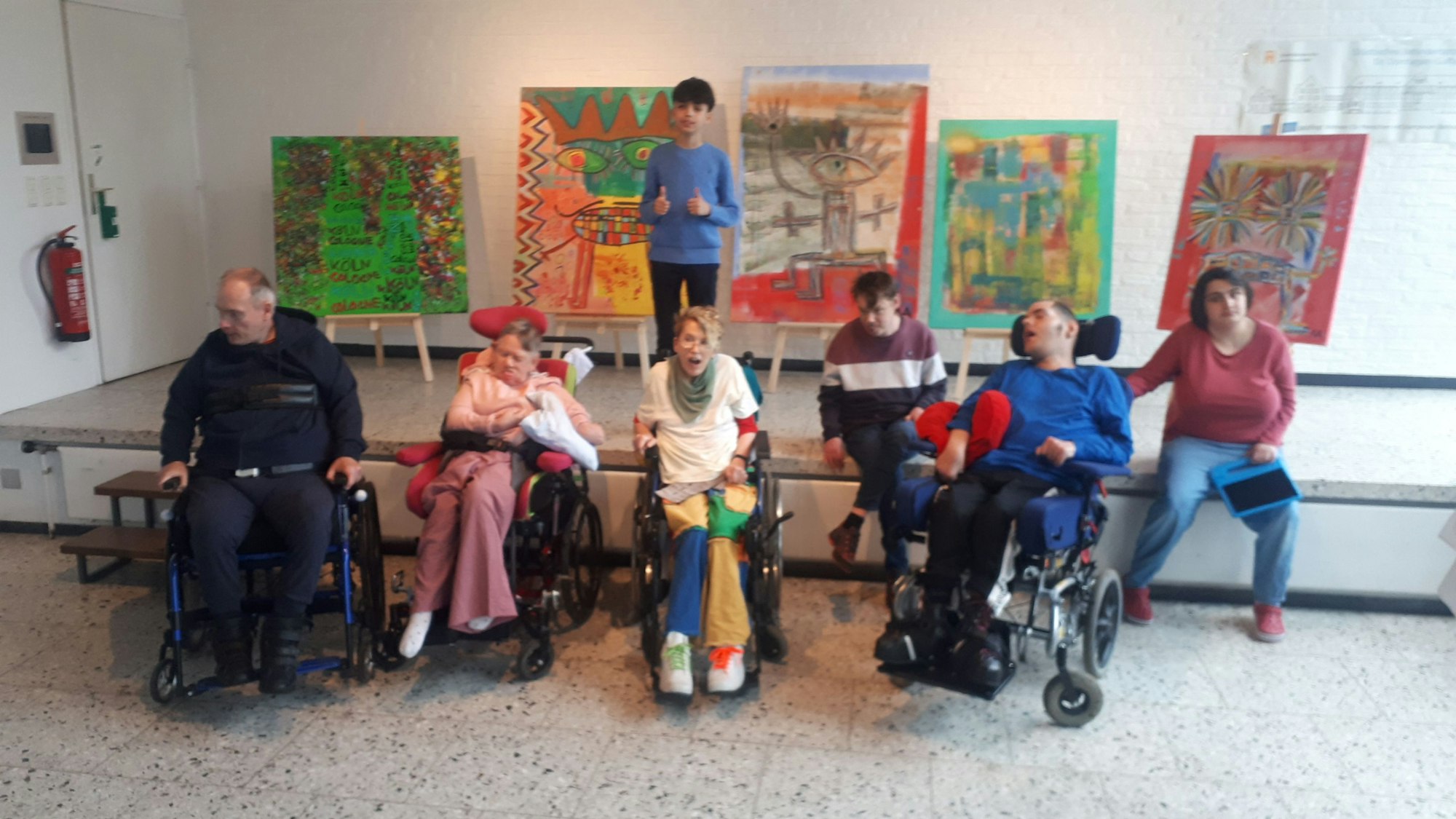 Menschen, teils im Rollstuhl sitzen vor einer Bühne, auf der fünf Bilder auf Staffleien stehen. Der Künstler, Mikail Akar, steht vor seinen Bildern.