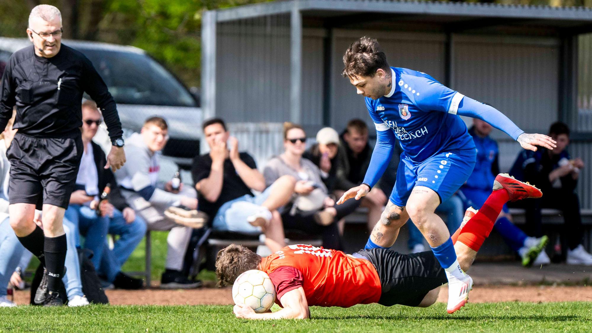 Ein Fußballer fällt nach einem Zweikampf auf den Boden und greift nach dem Ball.
