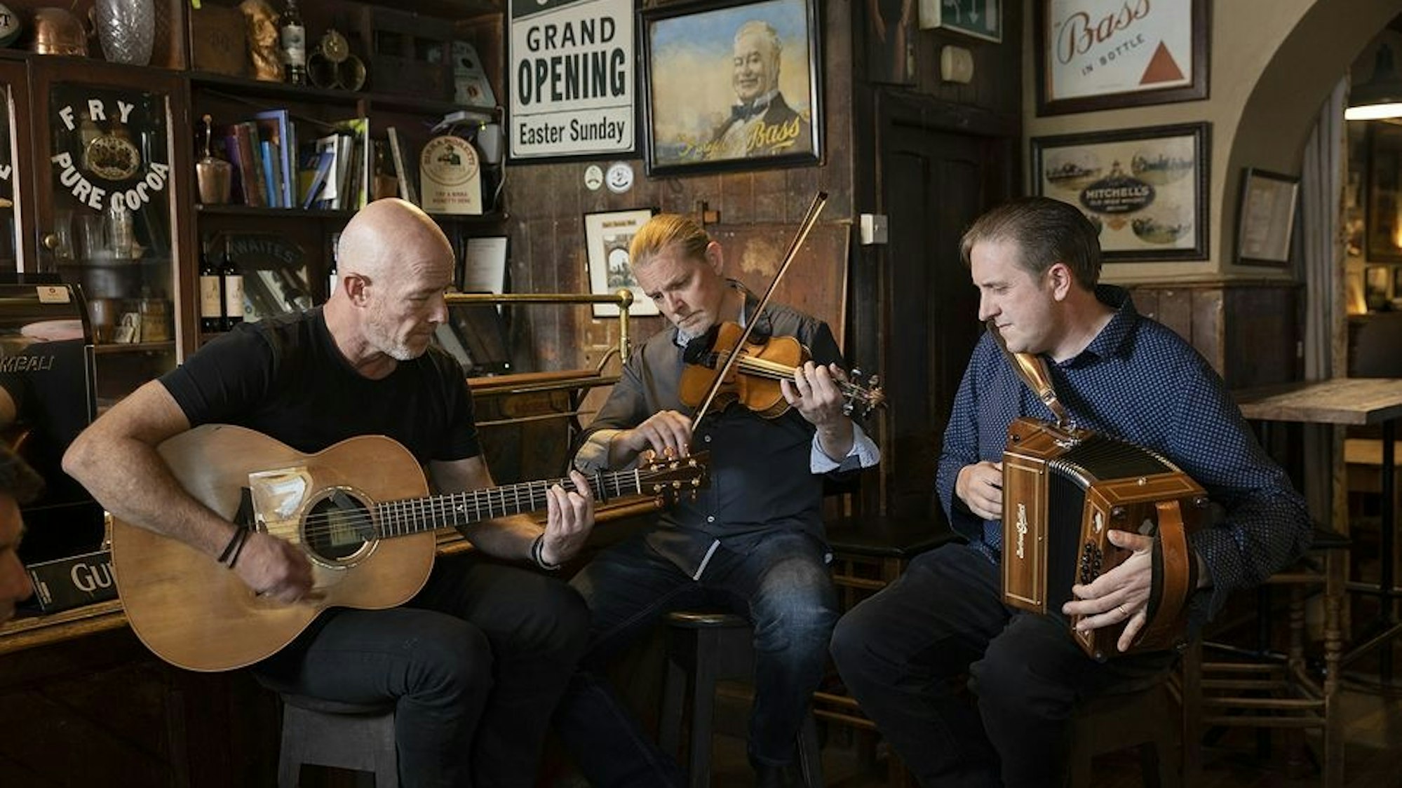 Drei Männer spielen Gitarre, Geige und Akkordeon in einem Pub.