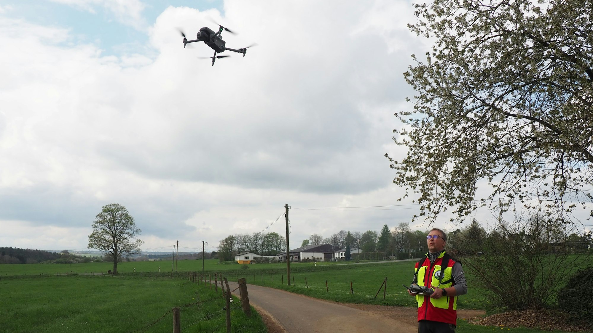 Ein Mann in rot-gelber Warnweste steht auf einem Weg. Er hält einen Controller in der Hand, mit dem er eine Drohne steuert, die links im Bild in der Luft zu sehen ist.