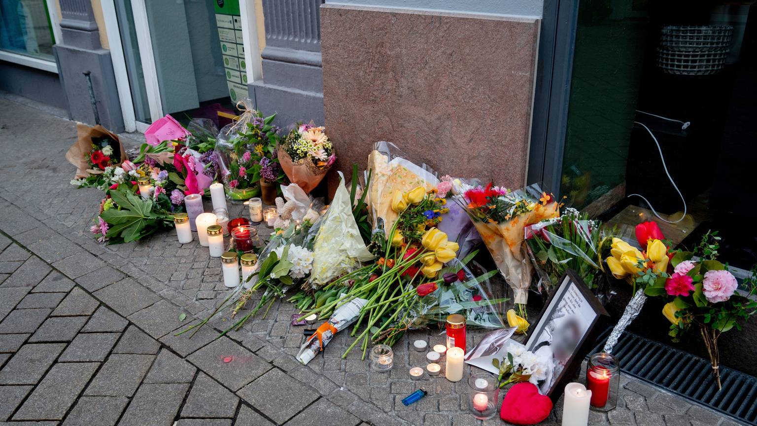 20.04.2025, Niedersachsen, Oldenburg: Zahlreiche Blumen und Kerzen stehen in der Innenstadt an einer Hauswand in der Achternstraße, in der am frühen Sonntagmorgen ein Mensch von Schüssen aus einer Polizeiwaffe tödlich verletzt wurde. Nach einer Auseinandersetzung in einem Lokal in der Innenstadt war der mutmaßliche Angreifer von Schüssen aus einer Polizeiwaffe tödlich verletzt worden. Foto: Hauke-Christian Dittrich/dpa - ACHTUNG: Foto des Toten gepixelt +++ dpa-Bildfunk +++