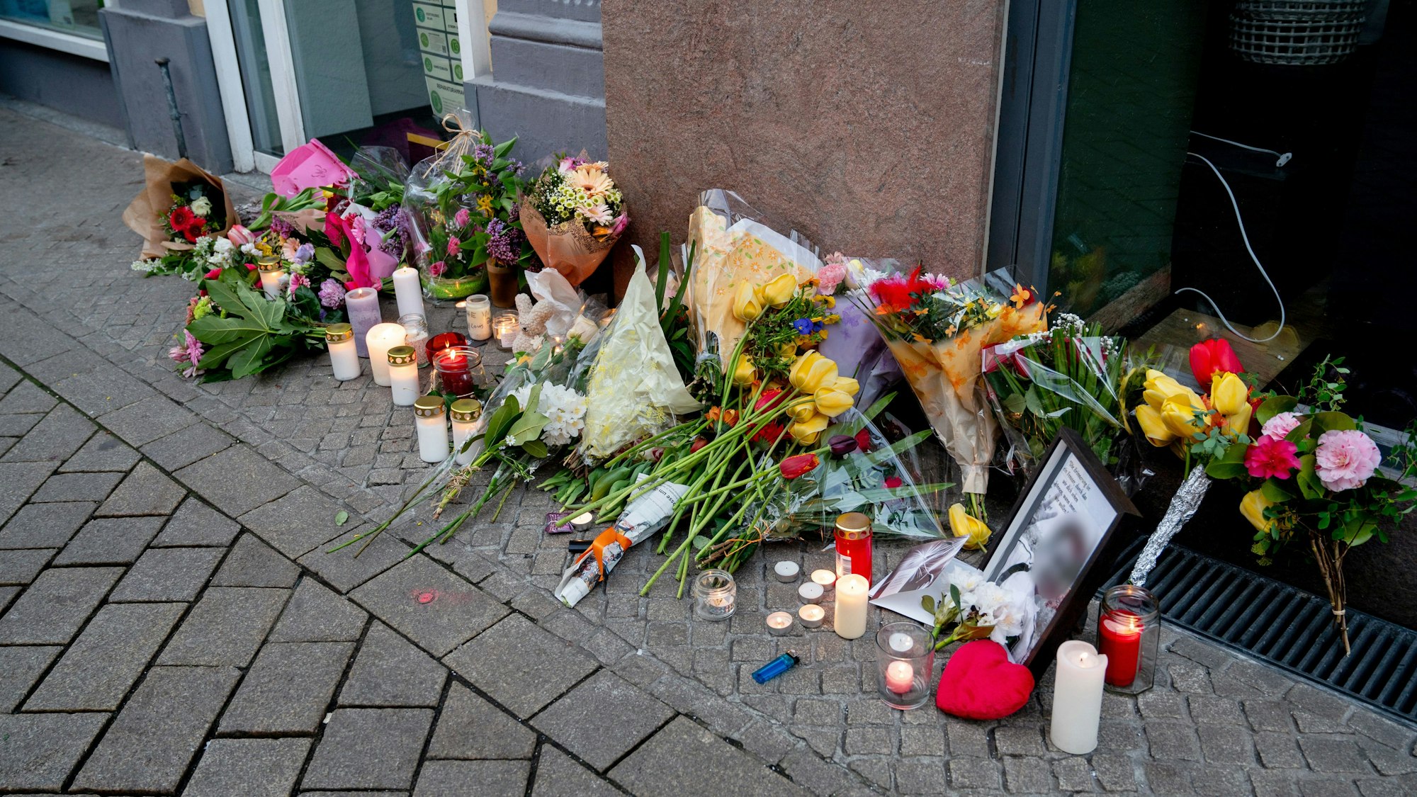 20.04.2025, Niedersachsen, Oldenburg: Zahlreiche Blumen und Kerzen stehen in der Innenstadt an einer Hauswand in der Achternstraße, in der am frühen Sonntagmorgen ein Mensch von Schüssen aus einer Polizeiwaffe tödlich verletzt wurde. Nach einer Auseinandersetzung in einem Lokal in der Innenstadt war der mutmaßliche Angreifer von Schüssen aus einer Polizeiwaffe tödlich verletzt worden. Foto: Hauke-Christian Dittrich/dpa - ACHTUNG: Foto des Toten gepixelt +++ dpa-Bildfunk +++