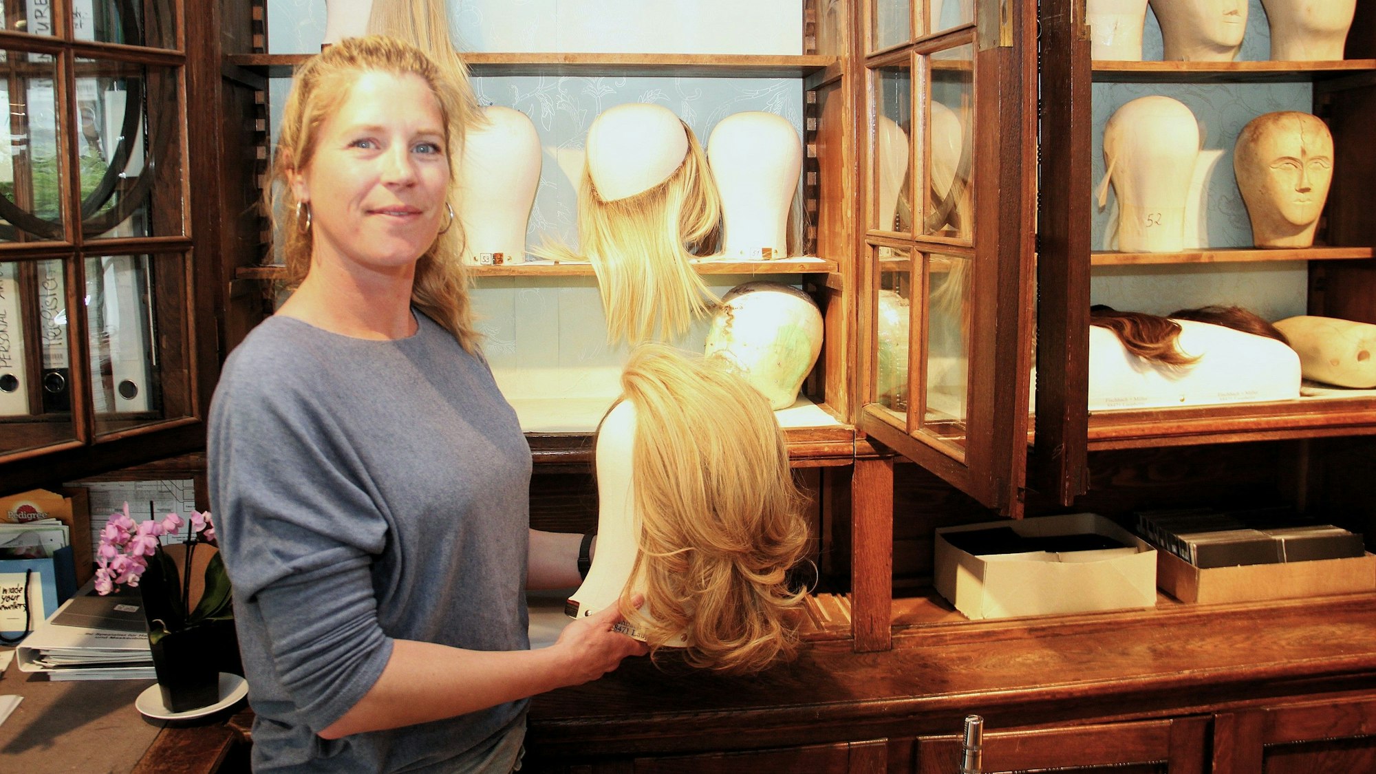 Eine Frau hält einen Perückenkopf mit Perücke in der Hand. Im Hintergrund stehen weiter Perücken in einem Regal.