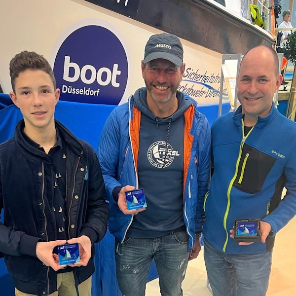 Zwei Männer und ein Jugendlicher stehen vor einem Schild der Boot in Düsseldorf und lächeln in die Kamera.