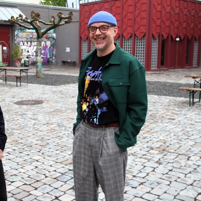 Eine Frau mit Locken und ein Mann mit Brille und blauer Mütze stehen in einem Innenhof.