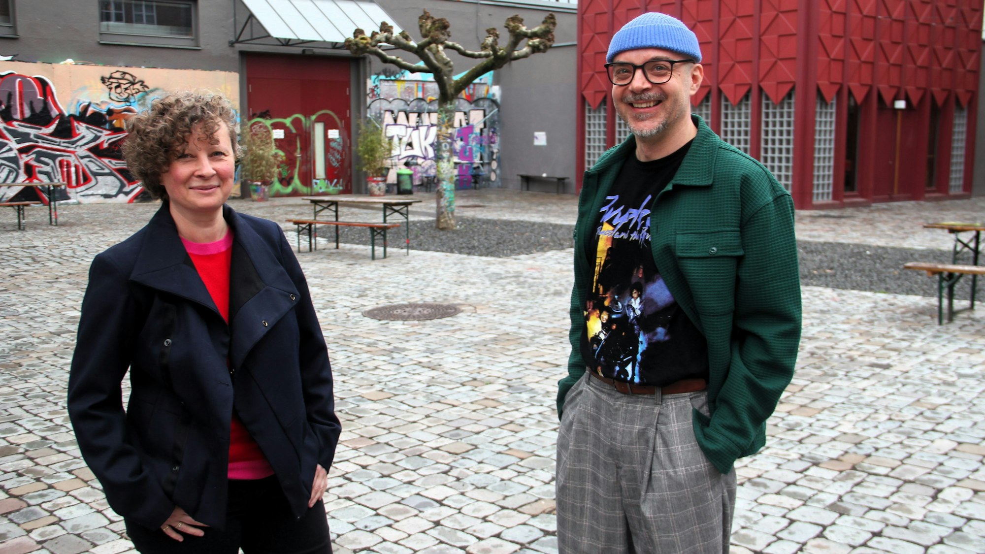 Eine Frau mit Locken und ein Mann mit Brille und blauer Mütze stehen in einem Innenhof.