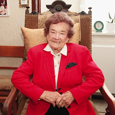 Eine betagte Seniorin im roten Jackett sitzt lächelnd in einem Sessel.