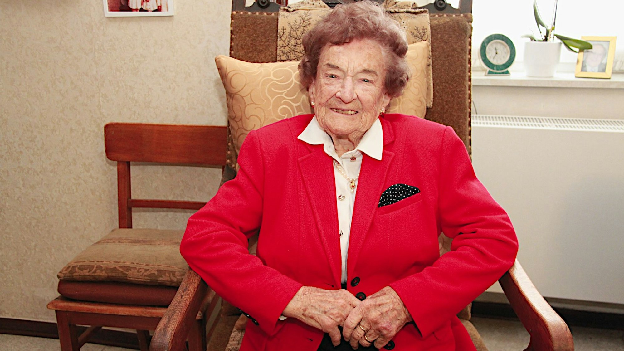 Eine betagte Seniorin im roten Jackett sitzt lächelnd in einem Sessel.