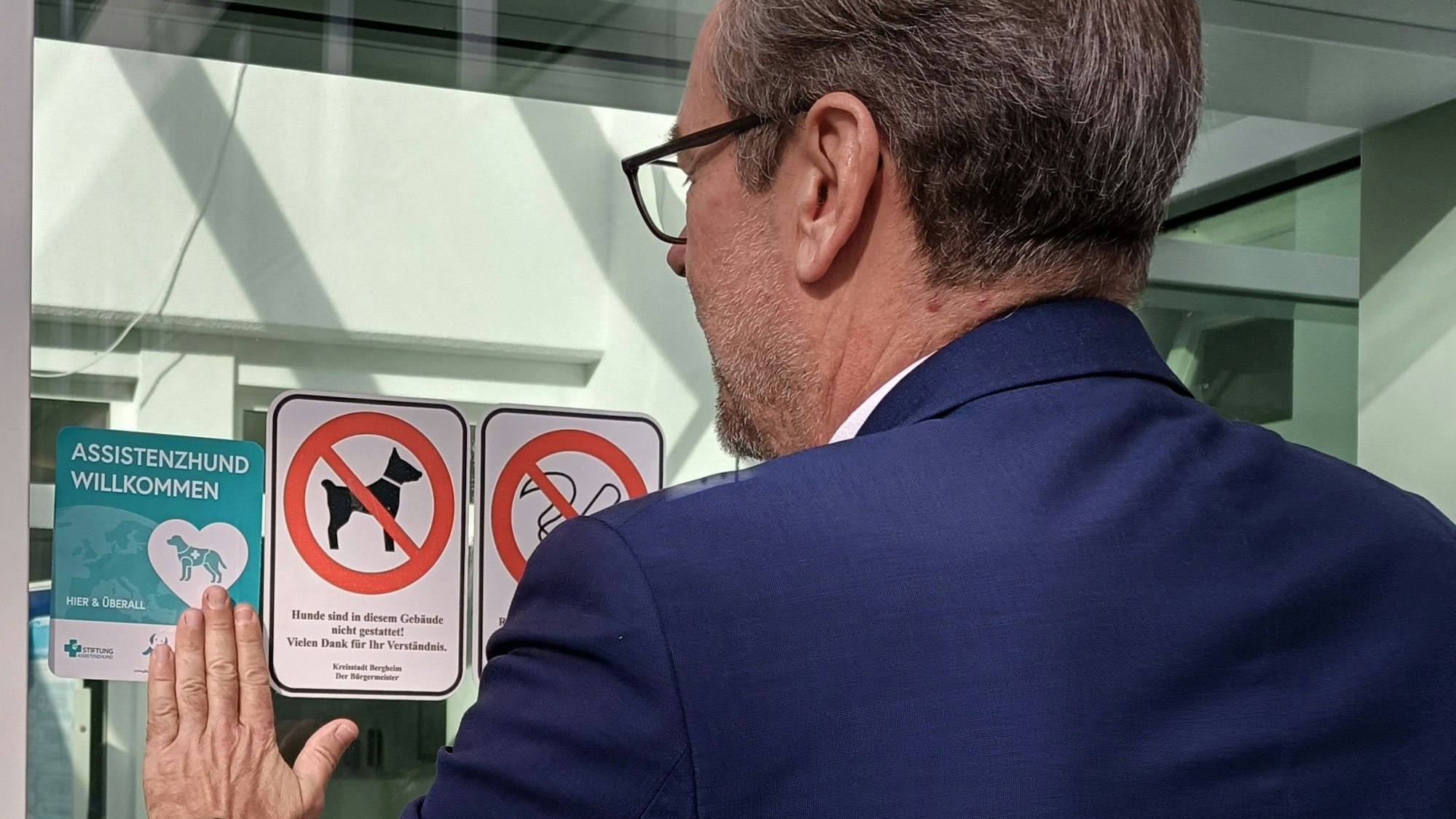 Ein Mann befestigt einen Aufkleber mit der Aufschrift „Assistenzhund willkommen“ an eine Glasscheibe.