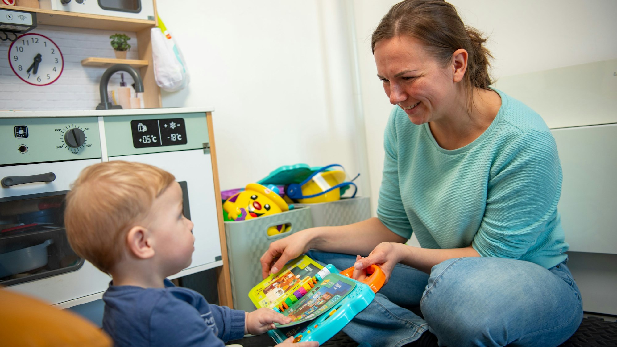 Eine Frau in einem türkisblauen Pullover und Jeans sitzt auf dem Boden und spielt mit einem Kind.