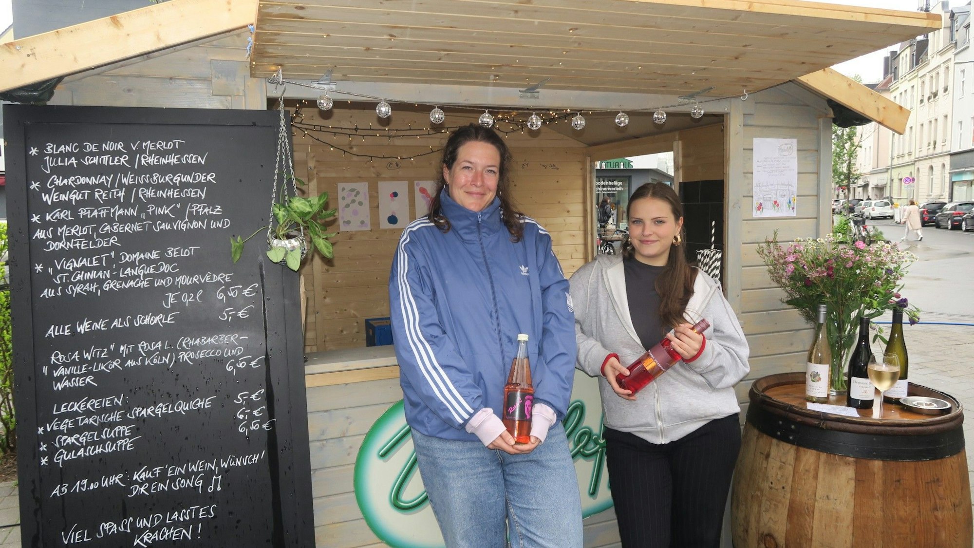 Zwei Frauen stehen unter dem Vordach einer kleinen Holz-Verkaufsbude, beide halten eine Flasche in den Händen.