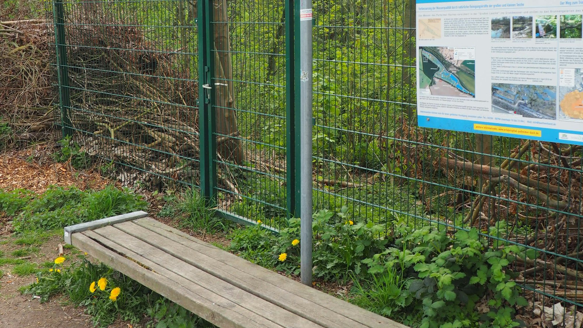 Im Vordergrund eine Bank, dahinter ein Zaun, an dem eine Infotafel hängt.