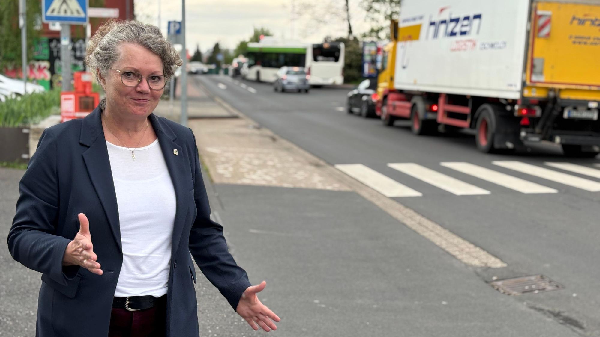 Bürgermeisterin Anna-Katharina Horst steht an einer Straße, an der mehrere Pkw, sowie ein Bus und Lkw stehen. Mit den Händen macht sie eine ausladende Geste.