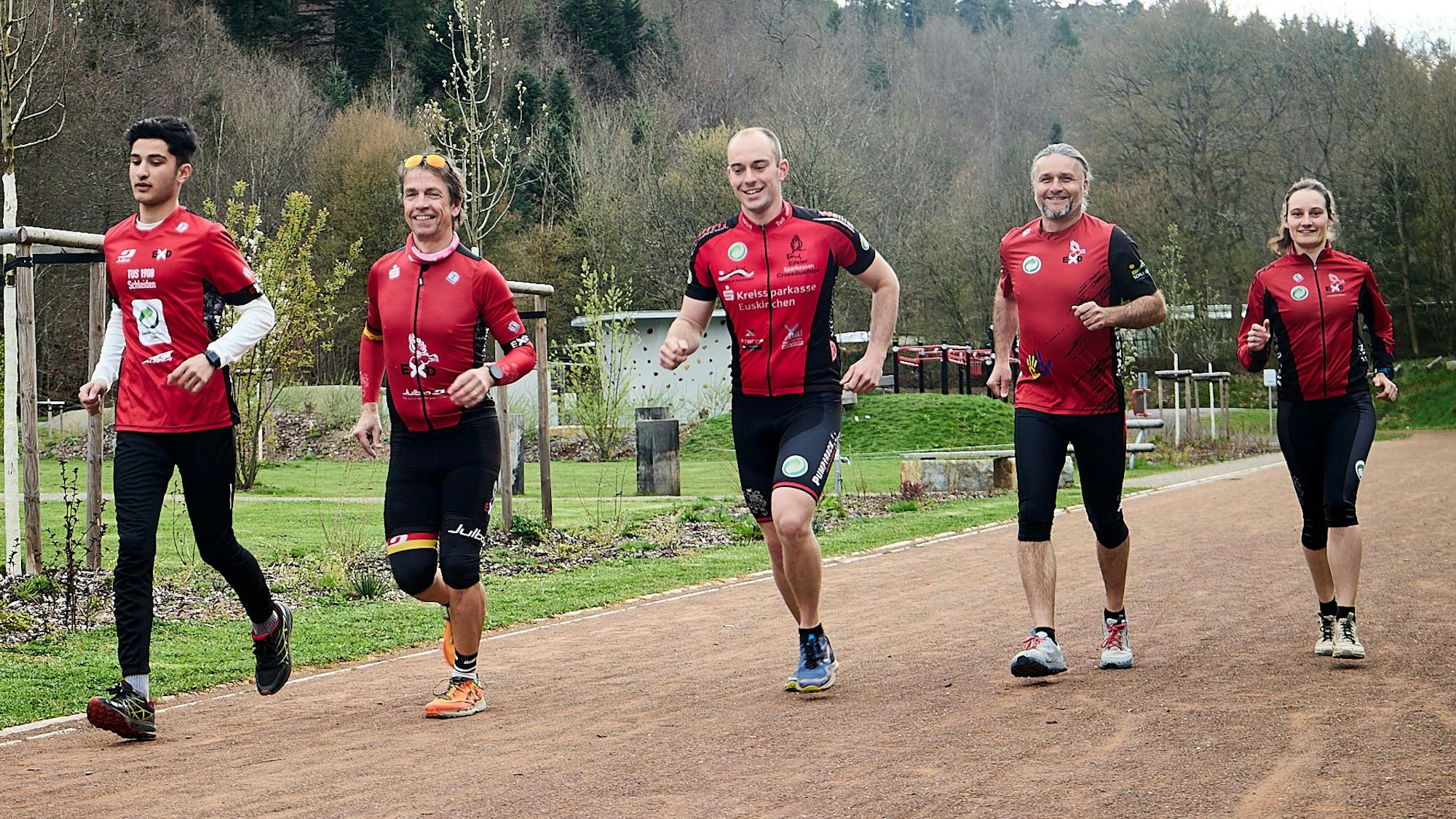 Vier Läufer und eine Läuferin in roten Trikots laufen auf einer Laufbahn eines Sportplatzes, im Hintergrund sind grüne Bäume zu erkennen.