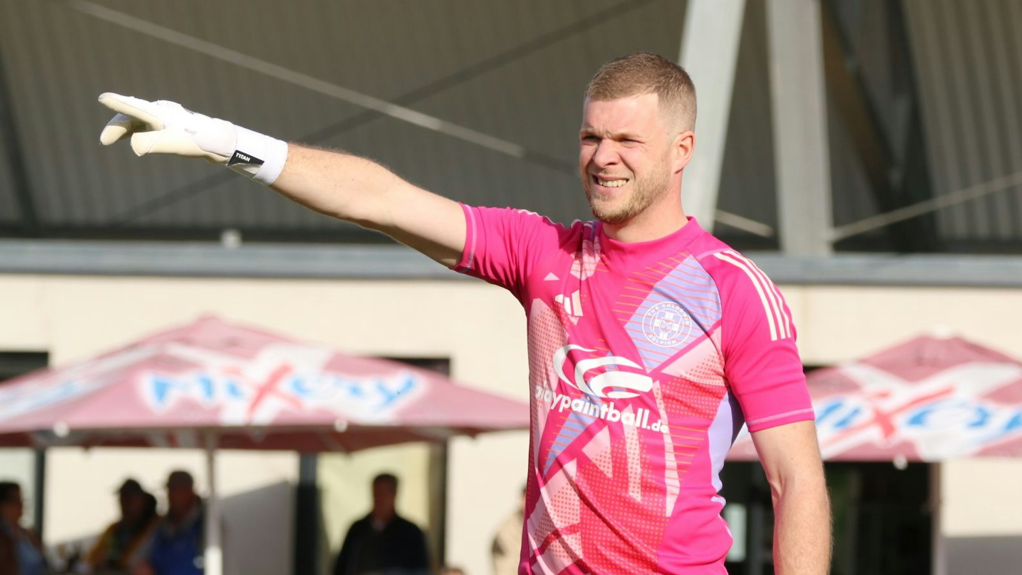 Robin Metternich trägt ein pinkes Torwarttrikot und weiße Torwarthandschuhe und hat den Zeigefinger der rechten Hand ausgestreckt.