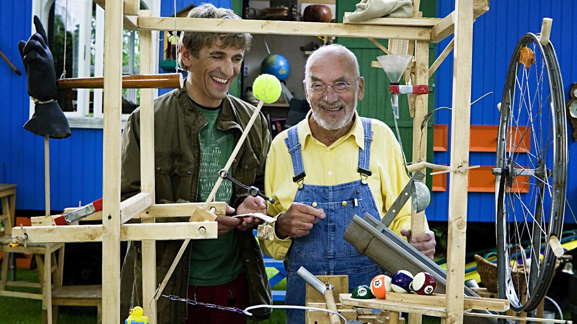 Fritz Fuchs (Guido Hammesfahr, l) und Peter Lustig stehen während einer Aufzeichnung für die ZDF-Kindersendung ‚Löwenzahn‘ hinter einer selbstgebauten Maschine.