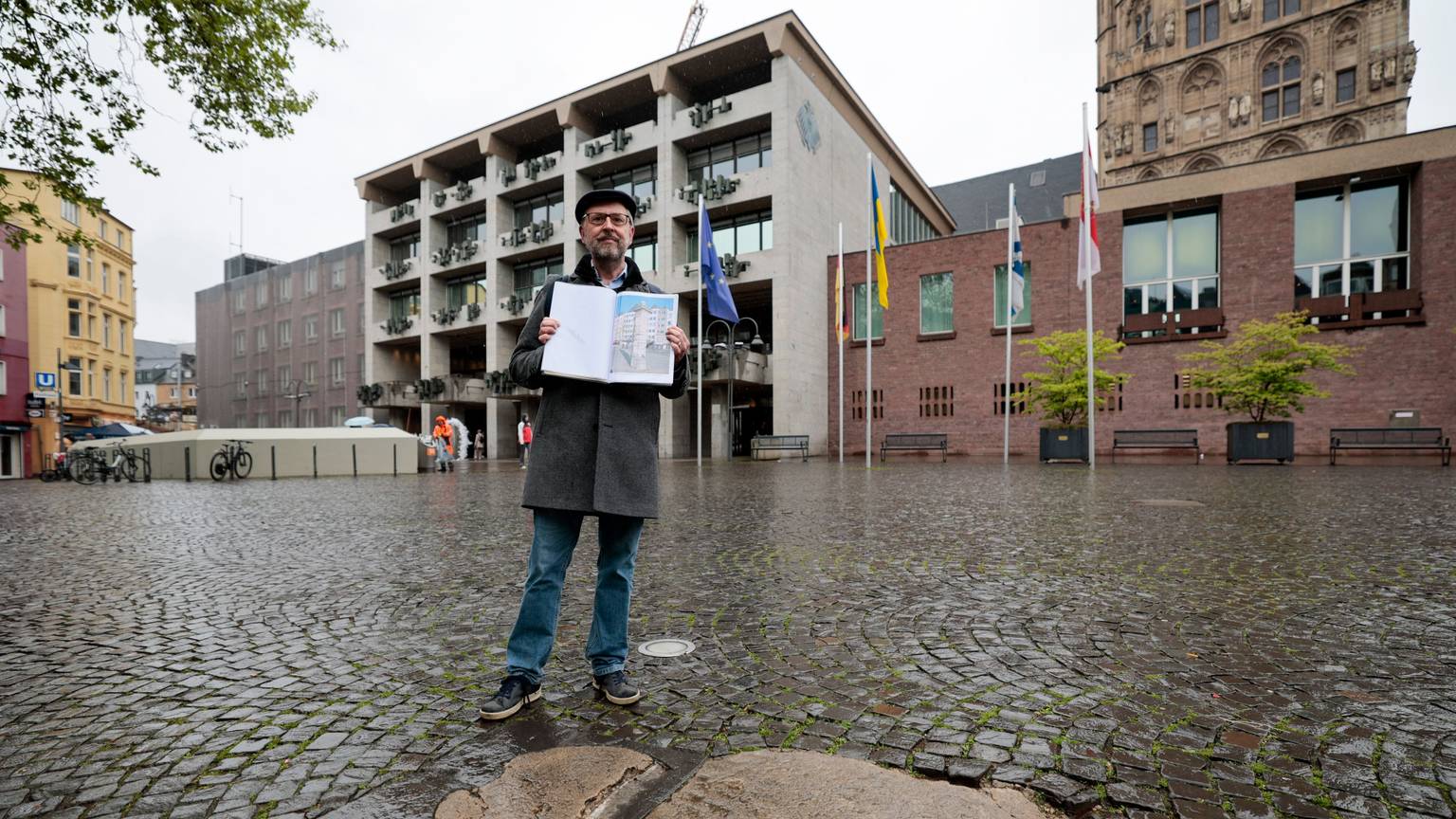 Ein Mann hält ein Fotoalbum mit Bilder der Historischen Wasserpumpe am Alter Markt hoch. Er steht vor dem Ort wo die Pumpe früher stand. Dort hebt sich ein heller Fleck vom restlichen Kopfsteinpflaster ab.
