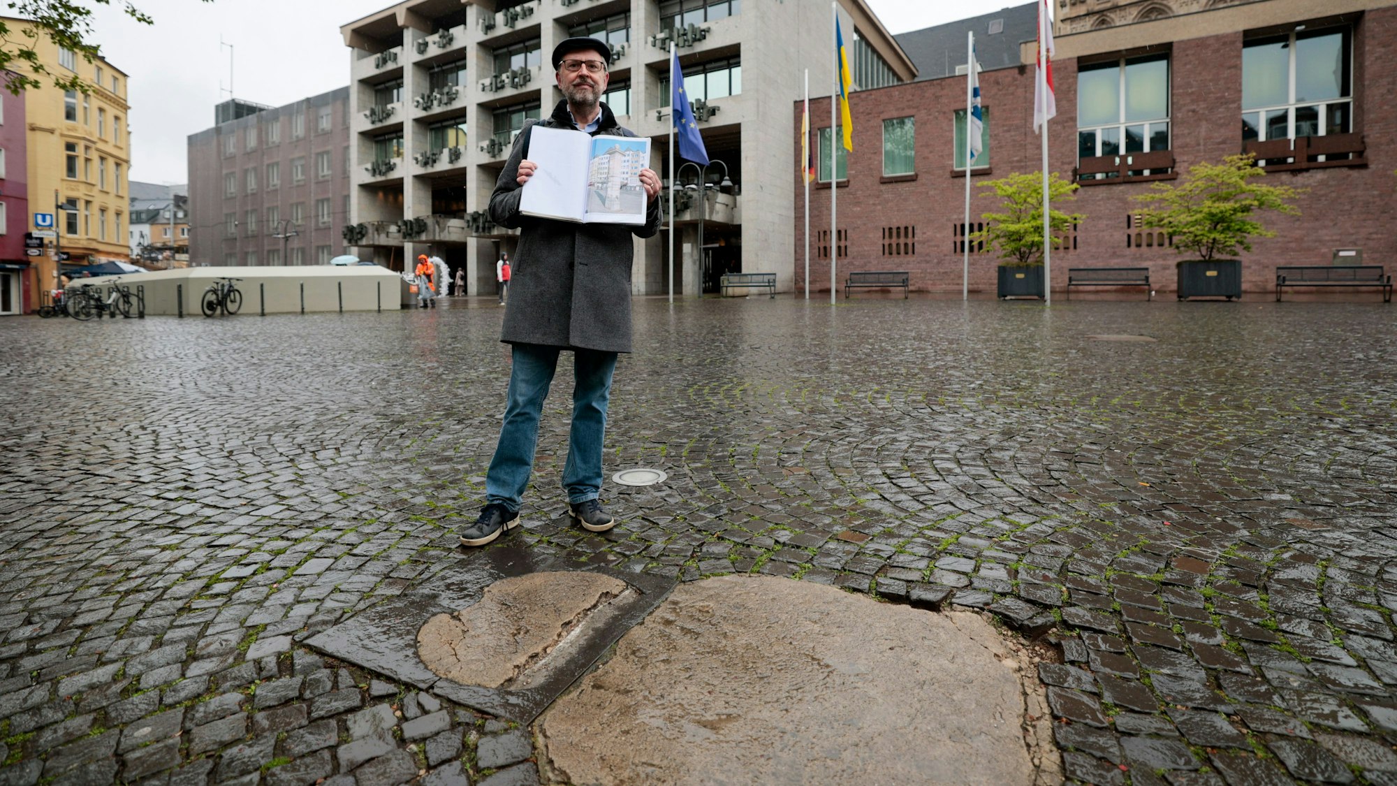 Ein Mann hält ein Fotoalbum mit Bilder der Historischen Wasserpumpe am Alter Markt hoch. Er steht vor dem Ort wo die Pumpe früher stand. Dort hebt sich ein heller Fleck vom restlichen Kopfsteinpflaster ab.