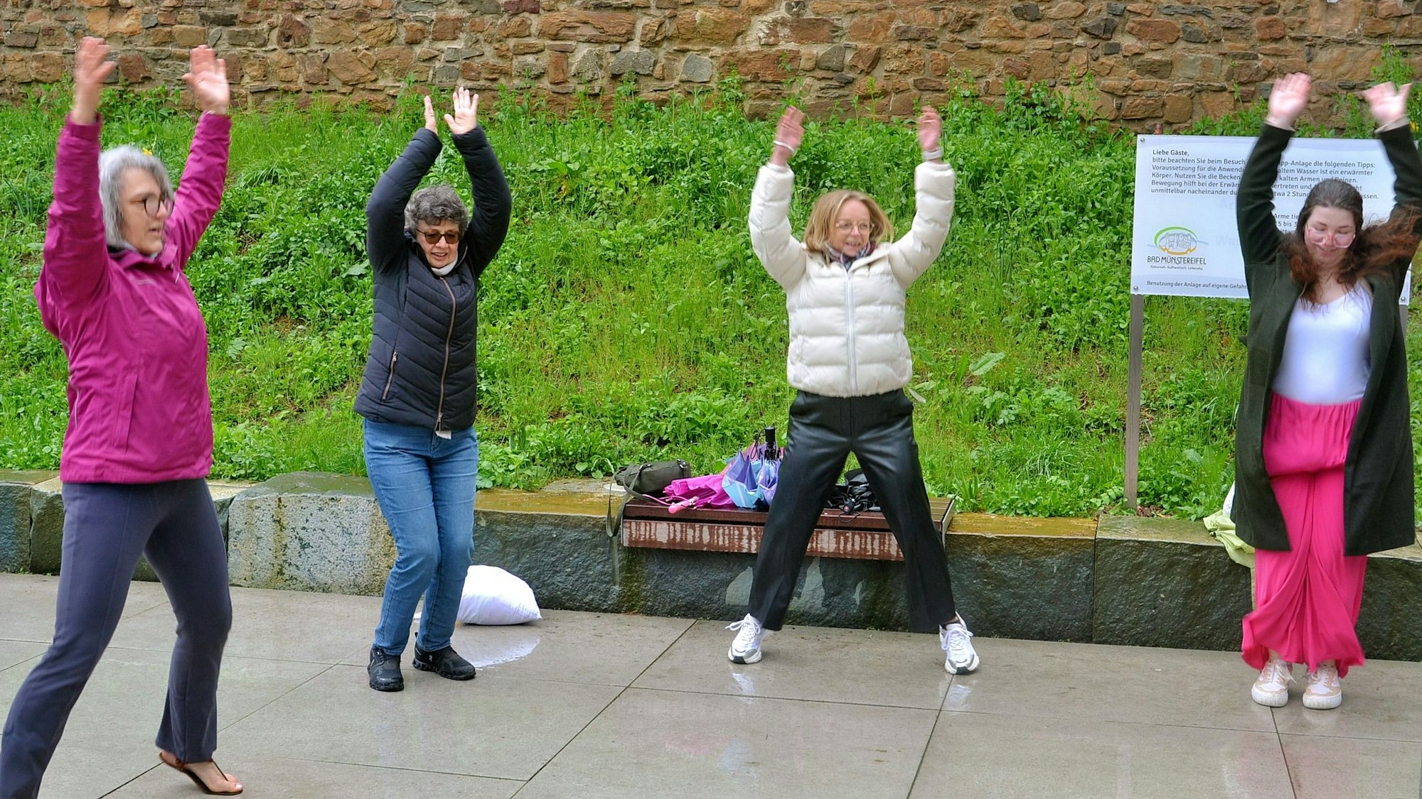 Vier Frauen stehen in Jacken und langen Hosen draußen in einem Halbkreis und machen Hampelmänner.