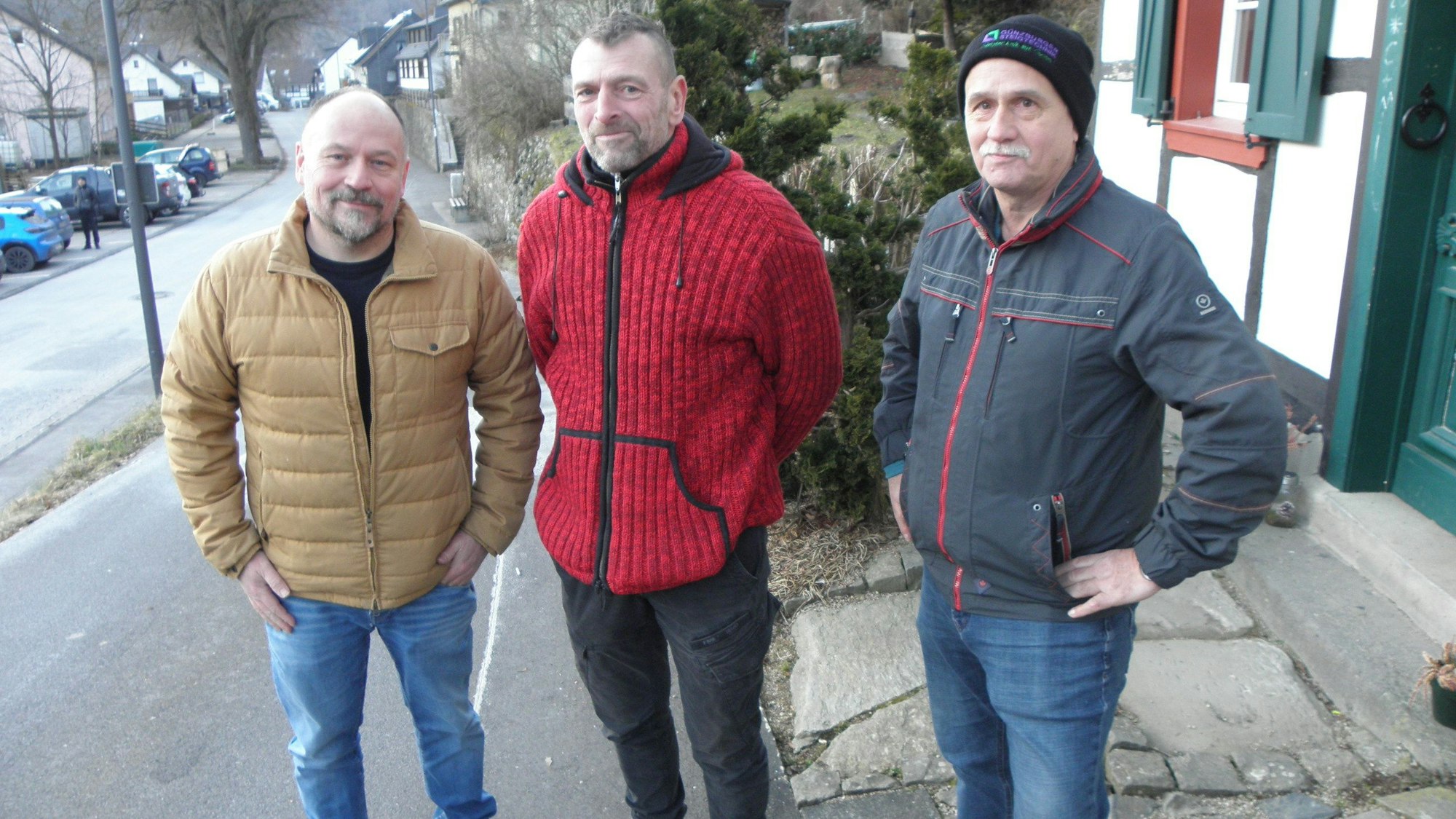 Drei Männer stehen in Jacken und Jeans an einer Straße.