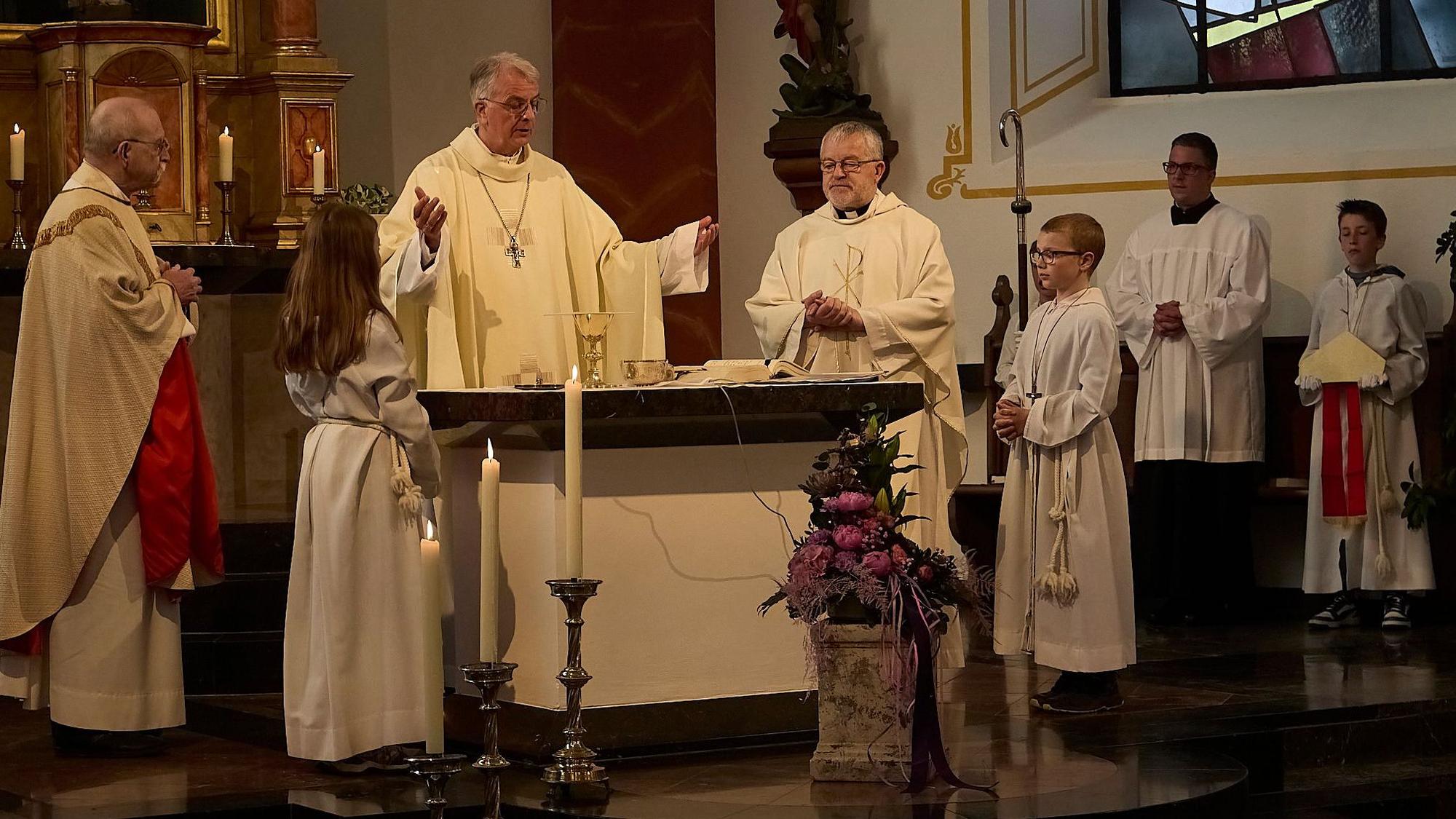 Der Trierer Weihbischof Jörg Michael Peters steht mit ausgebreiteten Armen am Alter in der Kirche in Nettersheim-Tondorf. Neben ihm stehen zwei weitere Priester und vier Messdiener.