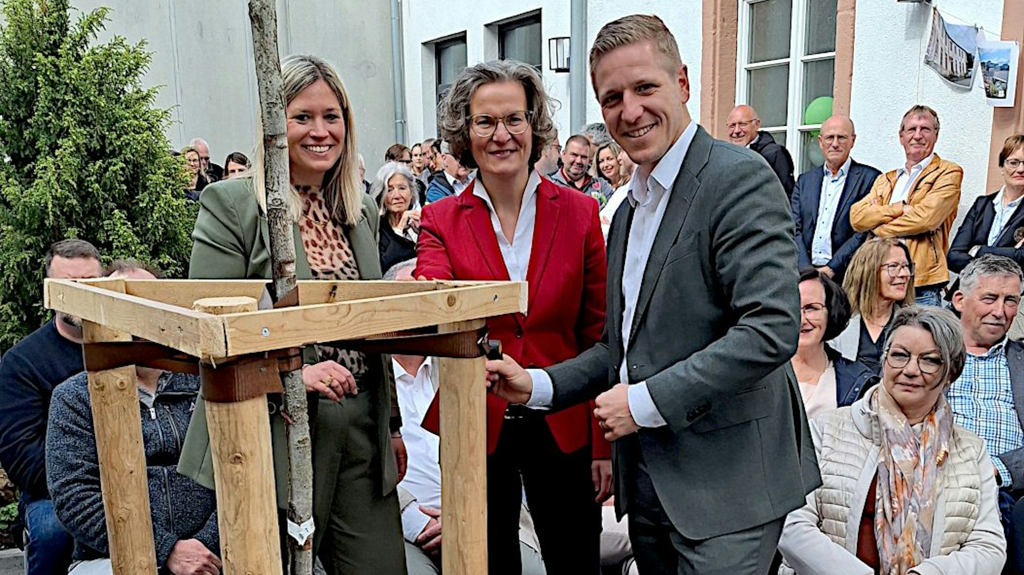 Jennifer Meuren, Ina Scharrenbach und Markus Ramers stehen um den Baum, dahinter sitzen und stehen Gäste der Feier.