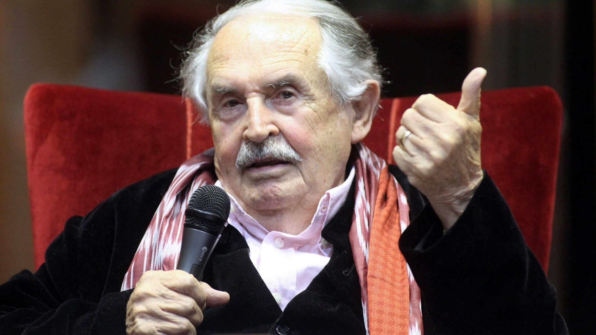 Ein alter Herr sitzt in einem Lehnsessel und spricht in ein Mikrofon.