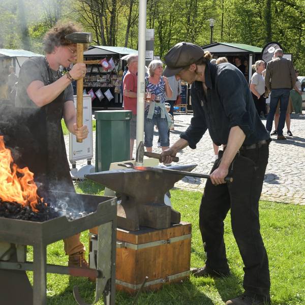 Zwei Personen hauen mit Hämmern auf eine Eisenstange, die auf einem Amboss liegt.