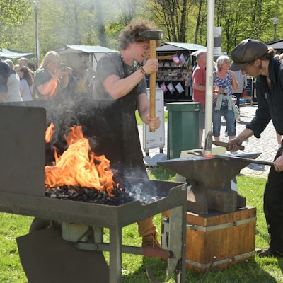 Zwei Personen hauen mit Hämmern auf eine Eisenstange, die auf einem Amboss liegt.